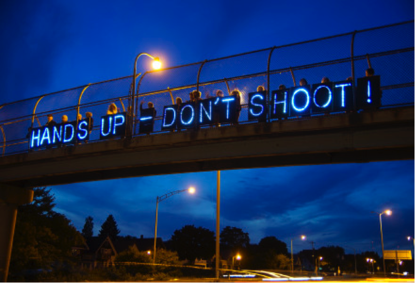 Image of light-up blue sign above a bridge reading: HANDS UP - DON’T SHOOT!