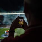 Person watching a football game from the stands taking a photo of the game with a smartphone.
