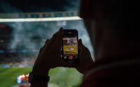 Person watching a football game from the stands taking a photo of the game with a smartphone.